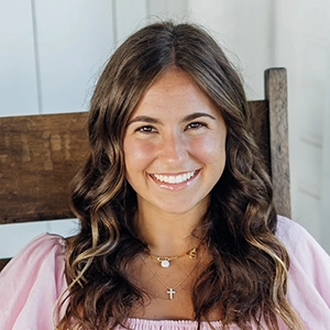 Smiling woman with long wavy brown hair, wearing a pink top and layered necklaces, seated indoors.