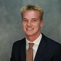 Studio headshot of a smiling young man in a dark suit and patterned tie against a gray background.