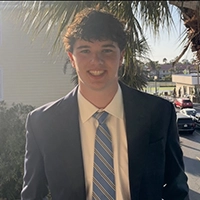 Smiling young man in a suit and striped tie standing outdoors near palm trees with buildings and parked cars in the background.