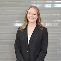 Professional headshot of a woman wearing a black blazer, standing against a light gray background.