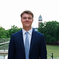 A young man in a dark suit and light tie stands confidently outdoors, with a blurred background featuring greenery and a tower.