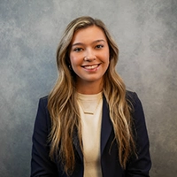 A professional headshot of a young woman with long blond hair, wearing a blazer and smiling against a neutral background.