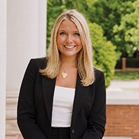 Smiling woman with blonde hair in a black blazer and white top, standing outdoors near a brick wall and columns.