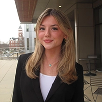 A young woman with blonde hair wearing a black blazer poses confidently outside a building.