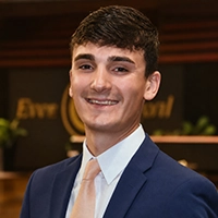 Headshot of a smiling young man in a navy suit and light tie, standing in an indoor office setting with a blurred logo in the background.