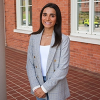 Smiling woman in a light gray blazer and white top standing with hands clasped in front of a brick building.