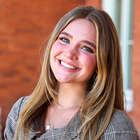 Smiling young woman with long light-brown hair standing in front of a brick building, looking at the camera.