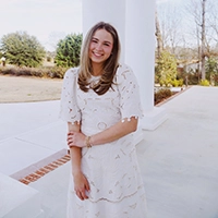 Smiling woman in a white embroidered dress leaning against a white column on a sunny outdoor walkway.