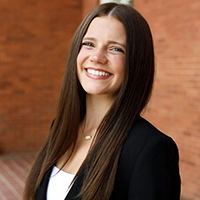 Smiling woman with long straight dark hair wearing a black blazer, posed in front of a brick wall.