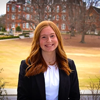 A smiling young woman in a professional outfit stands outdoors, with a university campus in the background.