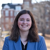 A young woman with shoulder-length brown hair smiles confidently while wearing a blue blazer. In the background, a blurred collegiate building is visible.