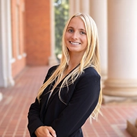 Smiling blonde woman in a black blazer standing with arms crossed in an outdoor brick colonnade.