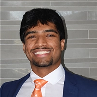 Smiling man in a navy suit with an orange tie standing in front of a gray tiled wall.