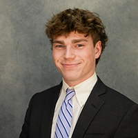 Smiling young man in a dark suit and striped tie against a gray studio backdrop.