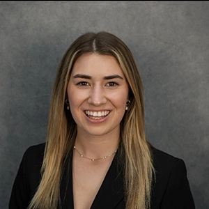 Smiling woman with long blond hair in a black blazer, posed against a gray studio backdrop.