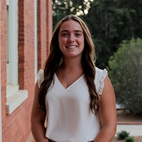 A smiling young woman with long hair stands outside near a brick building, wearing a white top.