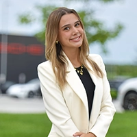 A young woman in a white blazer stands confidently outdoors, smiling at the camera.