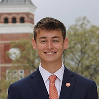 Smiling young man in a navy suit and orange tie, standing outdoors with a campus building and trees blurred in the background.
