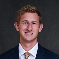 Head-and-shoulders studio portrait of a smiling person in a navy suit with a white shirt and patterned tie against a dark background.