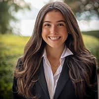 Smiling woman with long dark hair wearing a black blazer and white collared shirt outdoors, with a softly blurred green background.