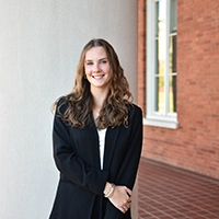 A smiling young woman standing in front of a brick building, wearing a black blazer.