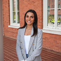 A smiling young woman in a gray plaid blazer stands in front of a brick building.