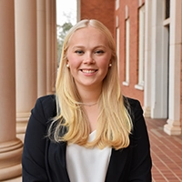 Professional portrait of a young woman with long blonde hair, wearing a black blazer and smiling confidently near classical columns.