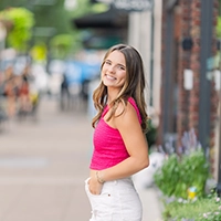 Smiling young woman in a pink top stands confidently on a vibrant street, showcasing a casual summer outfit.