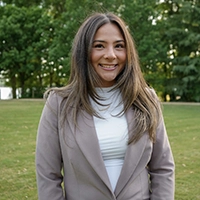 Smiling person with long brown hair wearing a beige blazer and white turtleneck, standing outdoors in a green park.