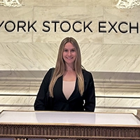 A woman stands in front of the New York Stock Exchange, smiling confidently.
