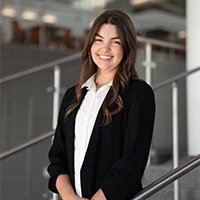 Smiling woman in a black blazer and white blouse standing indoors near a staircase railing.