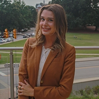 Smiling person in a brown blazer standing outdoors near a railing, with a road and green park in the background.