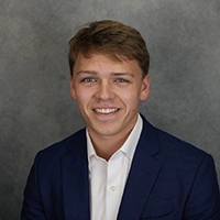 Smiling young man in a navy suit jacket and white shirt against a gray studio backdrop.