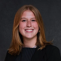 Smiling person with shoulder-length light brown hair, wearing a dark top against a dark studio background.