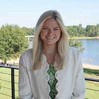 Smiling woman with long blonde hair wearing a white cardigan over a green patterned top, standing by a railing with a lake and trees in the background.