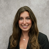 Professional headshot of a smiling woman with long brown hair wearing a black blazer against a gray backdrop.