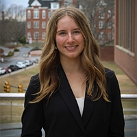 Smiling woman in a black blazer standing outdoors on a campus walkway, with brick buildings and parked cars in the background.