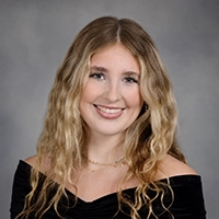 Studio portrait of a smiling person with long wavy blonde hair, wearing an off-the-shoulder black top against a gray background.