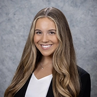 Professional headshot of a young woman with long, wavy hair, smiling and wearing a black blazer over a white top.