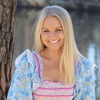 Smiling blonde woman outdoors by a tree with water in the background, wearing a blue floral top over a pink striped shirt.