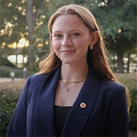 Smiling person in a navy blazer with a lapel pin, standing outdoors with trees softly blurred in the background.