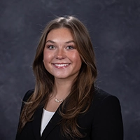 Professional headshot of a smiling woman with long brown hair, wearing a black blazer and a white top.