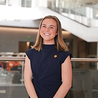A young woman with shoulder-length hair smiles confidently while wearing a dark blue top, standing in a modern building interior.