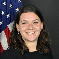 Headshot of a smiling woman with dark hair, wearing a black top, set against a backdrop of the American flag.