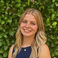 A smiling young woman with long blonde hair stands in front of a lush green background.