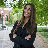 Smiling woman in a black blazer with arms crossed, standing on a tree-lined brick campus walkway with an arched building in the background.