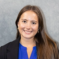 Smiling woman with long brown hair wearing a black blazer over a bright blue top against a gray studio backdrop.