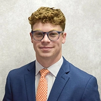 A professional headshot of a young man in a suit and tie, wearing glasses and smiling confidently.
