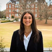 Smiling person in a black blazer and white shirt standing outdoors on a campus lawn with a brick building in the background.