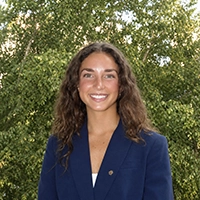 Smiling person in a navy blazer standing outdoors with leafy green trees in the background.